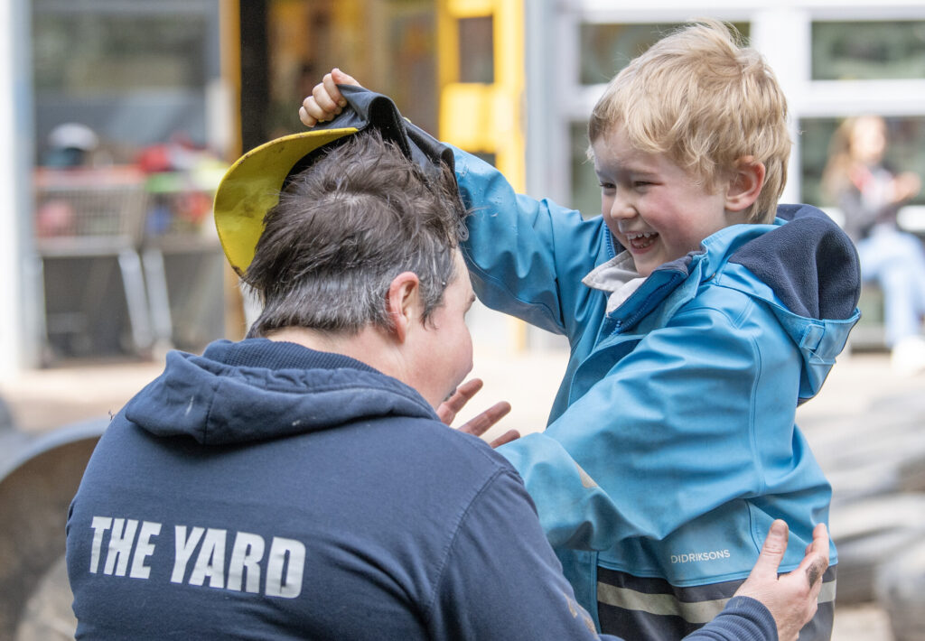 Playworker with little boy, he is taking her hat off and laughing