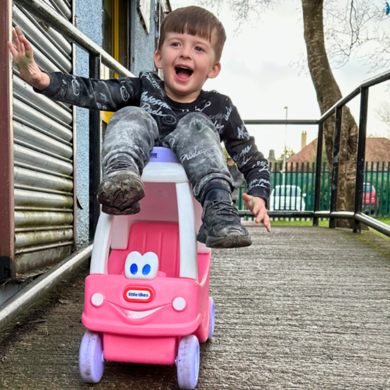 Little boy sitting on the roof on a toy pink car going downhill, smiling and laughing.