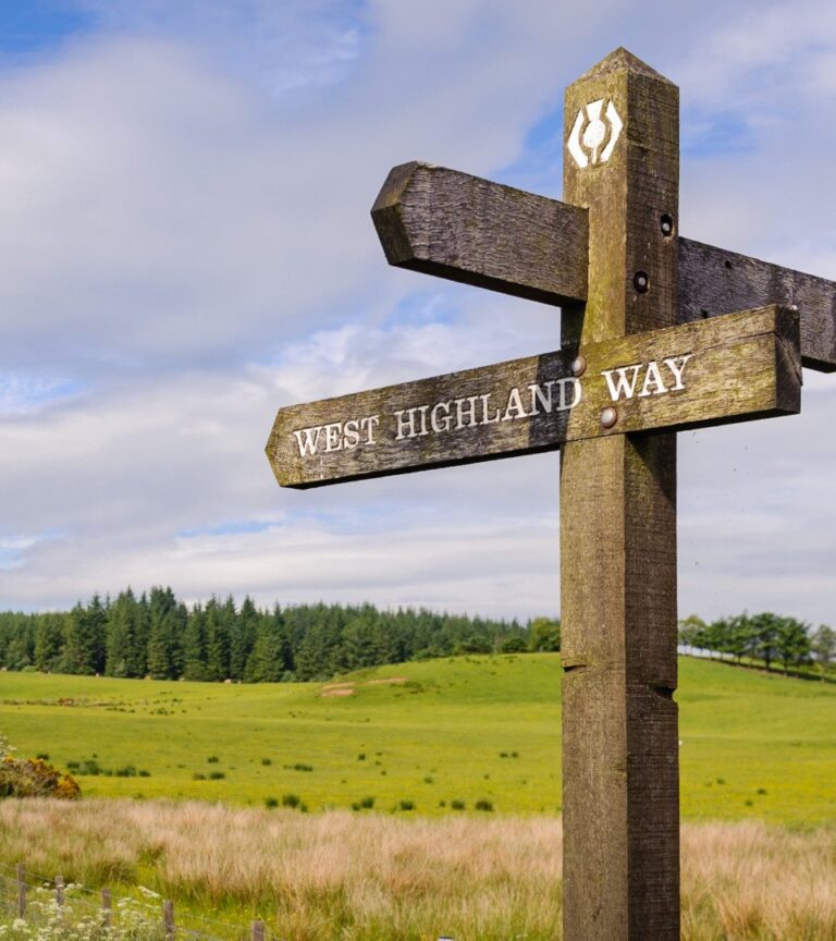 Signpost that says 'West Highland Way' with fields and hills in the background